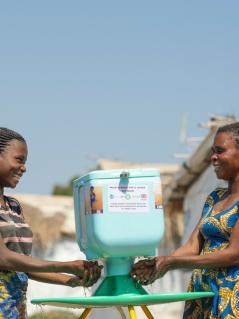 Zwei Frauen waschen ihre Hände an einer Handwasch-Station im Geflüchtetencamp in Kisalaba (Demokratische Republik Kongo). Die Frauen schauen einander lächelnd an.
