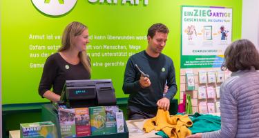 Zwei Ehrenamtliche stehen an einer Kasse in einem Oxfam-Shop und kassieren die Kleidung einer Kundin ab
