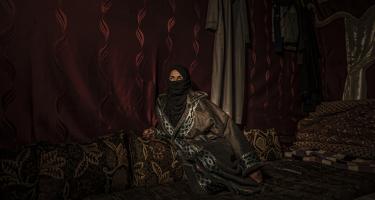 Eine Frau mit langen Gewändern sitzt vor einem roten Vorhang. Durch den Hijab sind nur ihre Augen zu sehen, die interessiert zur Seite schauen.
