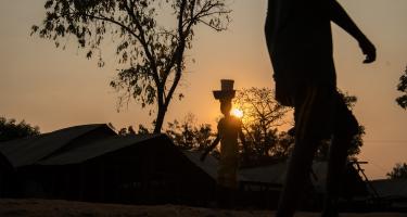 Ein Mensch trägt im Licht der Dämmerung zwei Gegenstände auf seinem Kopf. Der Sonnenuntergang legt sich warm über die Szenerie, ein zweiter Mensch läuft durchs Bild, aber auch dieser Mensch ist nur in seinen Umrissen erkennbar.