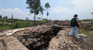 Auf einer flachen, grünen Landschaft sind die Überreste einer zerstörten Latrinenanlage von Oxfam zu sehen. An der Stelle der Latrinen ist nur noch eine tiefe Steingrube übrig. Eine Person, die für Oxfam arbeitet, steht rechts neben der verwüsteten Stelle und sammelt Überreste auf.
