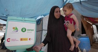 Duaa Abu Sabha benutzt eine Handwaschstation, die Oxfam für Geflüchtete im Gebiet Al-Mawasi im Gouvernement Khan Yunis aufgestellt hat. Duaa Abu Sabha wäscht sich die Hände an einer Handwaschstation, die in einem Camp für Geflüchtete steht. Auf dem Arm trägt sie ein Kleinkind.