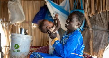 „Als Geflüchtete im Südsudan erhalte ich eine monatliche finanzielle Unterstützung, Hygieneartikel und Seife von Oxfam. Das hilft mir, aber ich würde gerne mehr für die Ernährung meiner Kinder tun.“ Ayul Ajak, Mutter von sechs Kindern und sehbehindert, ist vor dem Krieg im Sudan geflohen. Ayul Ajak sitzt in einer provisorischen Unterkunft und spielt mit einem Kleinkind. Im Hintergrund steht ein Eimer mit der Aufschrift „Oxfam“.