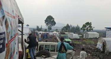 In einem vom diesigen Nebel der Regenzeit umgebenen Camp steht eine Gruppe von Menschen zwischen feuchten Zelten, während ein LKW mit weiteren Zeltplanen ankommt; ein Geländewagen parkt im Matsch.