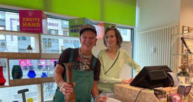 Ein Mann und eine Frau stehen lächelnd hinter der Kasse eines Oxfam Shops. Der Mann hält eine Vase in der Hand. 
