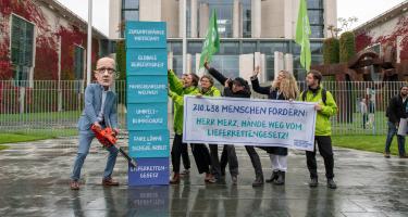Eine Gruppe mit Oxfam-Fahnen vor dem Kanzleramt. Auf ihrem Transparent steht: „210438 Menschen fordern: Herr Merz, Hände weg vom Lieferkettengesetz!“ Eine Person mit Friedrich-Merz-Maske sägt mit einer Kettensäge an einem symbolischen Turm. Der unterste Baustein des Turms ist mit „Lieferkettengesetz“ beschriftet, darüber aufgebaut sind: faire Löhne und sichere Arbeit, Umwelt- und Klimaschutz, Menschenrechte weltweit, globale Gerechtigkeit und zukunftsfähige Wirtschaft.