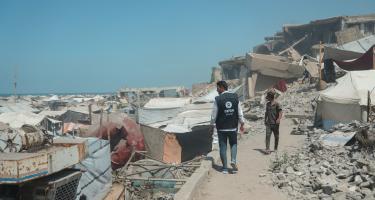 Oxfam-Mitarbeiter Osama Qatta zwischen Zelten und Trümmern in Gaza-Stadt. Ein Mann in Oxfam-Weste läuft auf einem schmalen Weg zwischen Trümmern, Steinen, Metallteilen und Zelten. Ein unbekannter Mann kommt ihm entgegen.