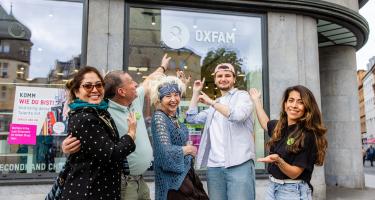 Fünf Personen stehen draußen vor dem Oxfam Shop in Halle und zeigen fröhlich auf das Logo.