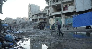Ein Mann schiebt einen Karren mit Kanistern durch die Trümmer in Gaza-Stadt. Der Weg ist überschwemmt mit Abwasser. Im Hintergrund Ruinen und überall Schutt.