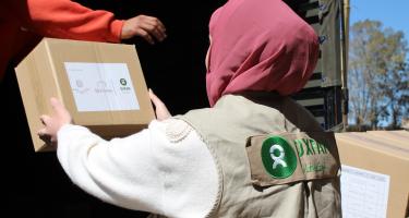 Eine Person mit beiger Oxfam-Weste und rosa Kopftuch lädt Nothilfe-Pakete aus einem Lkw.