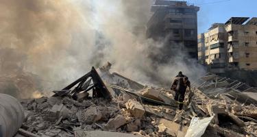 Die Trümmer eines Hauses zwischen anderen Gebäuden. Es steigt Rauch auf. Zwei Menschen bewegen sich über die Trümmer.