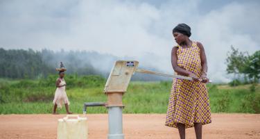 Eine Frau pumpt Wasser in einen Kanister. Hinter transportiert eine andere Frau ein Bündel auf dem Kopf. Im Hintergrund Grün und Nebel