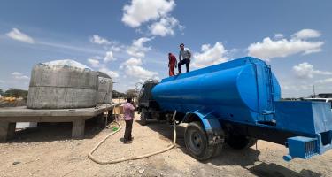Links im Bild stehen zwei große Betontanks auf einem Podest, rechts daneben ein blauer Tankwagen, der per Schlauch einen der Wassertanks füllt. In der Mitte und auf dem Tankwagen stehen drei Menschen.