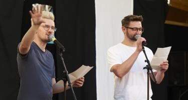 Zwei Männer sprechen in Mikrofone. Sie halten jeweils ein Blatt Papier in der Hand.