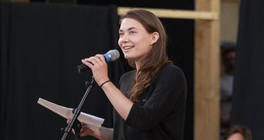 Eine Frau hält lächelnd ein Mikrofon in der einen Hand und Papierblätter in der anderen Hand.