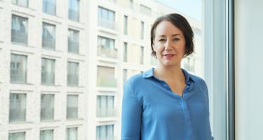 Eine Frau lehnt an einem großen Bürofenster und schaut in die Kamera. Im Hintergrund ein Bürogebäude.