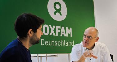 Zwei Männer im Gespräch. Sie sitzen vor einem Schild mit der Aufschrift „Oxfam Deutschland.“.