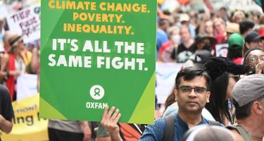 Ein Mann in einer großen Menge Demonstrierender. In der Hand hält er ein Plakat mit der Aufschrift „Climate Change. Poverty. Inequality. It's all the Same Fight.“ (Deutsch: „Klimawandel. Armut. Ungleichheit. Es ist alles derselbe Kampf.")