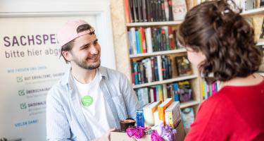 Eine Frau übergibt einem Mann einen Karton mit Büchern, Halstüchern und anderen Dingen.