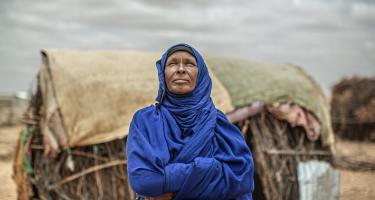 Amina Ibrahim schaut in einen wolkenbedeckten Himmel und verschränkt die Arme vor der Brust. Im Hintergrund ist ein einfaches Haus aus Decken und Holz zu sehen.