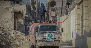 Ein Tanklastwagen in einer Straße in Aleppo. Auf dem Wagen steht ein Mann mit einem Schlauch in der Hand. Links neben dem Wagen sieht man die Ruinen eines Hauses.