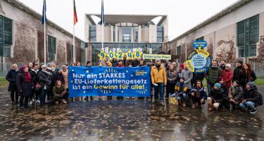 Aktivist*innen der "Initiative Lieferkettengesetzt" mit einem Banner "Nur ein starkes EU-Lieferkettengesetz ist ein Gewinn für alle!" vor dem Bundeskanzleramt. In der hinteren Reihe halten einige Aktivist*innen die Anzahl der eingereichten Unterschriften (90.248) hoch. Rechts ist ein Glücksrad zu sehen, das den "Gewinn für alle" als Hauptgewinn anzeigt.