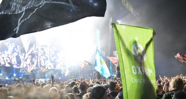Vor der Konzertbühne hält eine Konzert-Aktivistin, auf den Schultern eines anderen Aktivisten sitzend, die Oxfam-Fahne in die Luft. Sie sind umgeben von einer Menge an Fans.