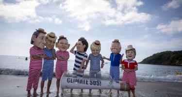 Aktivist*innen posieren als G7-Regierungschef*innen am Strand. In gestreiften Badesachen und Sonnenhüten stehen sie um eine riesige Flaschenpost herum und schauen sich fragend oder skeptisch an.