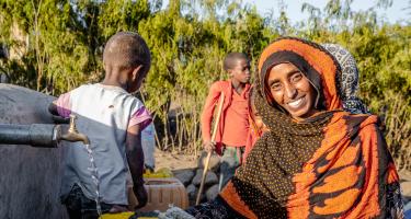 Kommt dank Wasserquelle besser durch den Alltag: Seadiya Mohamed Seadiya Mohamed lächelt in die Kamera, während sie Wasser aus einem laufenden Hahn in ihren Kanister schöpft. Im Hintergrund stehen zwei Kinder.