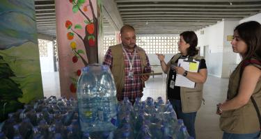 Drei Personen stehen hinter einem großen Stapel Wasserflaschen. Sie halten Klemmbretter in der Hand und sprechen miteinander.