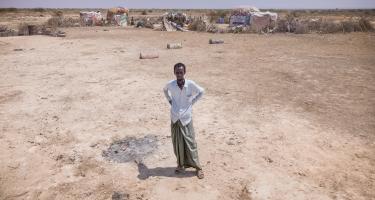 Abdulahi Farah Isse steht, bekleidet mit einem weißen Shirt und einem grauen Wickelrock, auf einem sehr trockenen Boden. Im Hintergrund ist seine Unterkunft zu sehen.