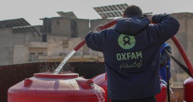 Ein_e Mitarbeiter_in von Oxfam in Syrien befüllt einen Wassertank, damit die Menschen in Notunterkünften Zugang zu sauberem Trinkwasser haben. Ein_e Oxfam-Mitarbeiter_in, mit einer blauen Kaputzenjacke mit grünem Oxfam-Logo bekleidet, befüllt mithilfe eines Schlauches einen roten Wassertank mit sauberem Trinkwasser.
