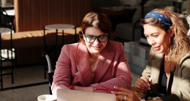Zwei Frauen sitzen an einem Holztisch und schauen gemeinsam auf einen Laptop