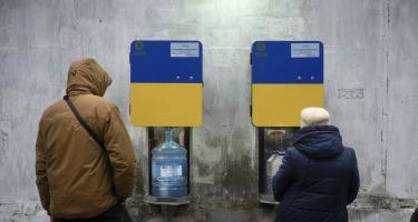 2 Menschen stehen vor zwei Trinkwasserautomaten, aus denen Wasser in ihre Flaschen läuft.