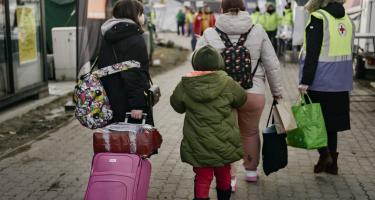 Eine Gruppe von Menschen mit Koffern, Rucksäcken und Taschen bepackt, läuft eine Straße entlang. Sie tragen dicke Kleidung und Masken. Sie werden von einer Person mit neongelber Weste mit rotem Kreuz-Symbol begleitet. Im Hintergrund sind Zelte und Menschen mit Warnwesten zu sehen.