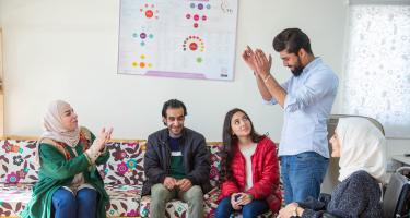 Hala, Rana, Abed und Mohammed sitzen in einem Kreis, ihr Geschäftspartner steht zwischen ihnen.