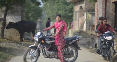 Eine junge indische Frau sitzt auf einem Motorrad; neben ihr zwei Männer auf einem anderen Motorrad
