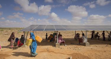 Wegen des Klimawandels haben die Menschen am Horn von Afrika immer häufiger mit Dürren zu kämpfen. Ein Mädchen trägt Wasserkanister, während eine Gruppe von Menschen im Schatten einer Solaranlage sitzt