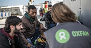 Eine Frau mit Oxfam-Weste, Rücken zur Kamera, spricht mit einer Familie in einem Flüchtlingscamp.