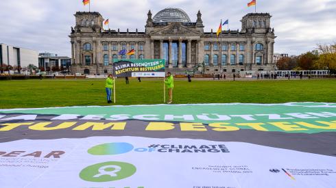Oxfam-Aktivist*innen, mit grünen Jacken bekleidet, stehen vor dem Reichstag und halten ein Banner mit der Aufschrift "Klima schützen - Reichtum besteuern" zwischen ihnen. Vor ihnen auf der Wiese befindet sich ein überdemensionales Transparent auf der Wiese.