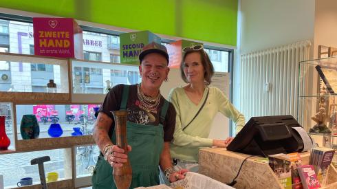 Ein Mann und eine Frau stehen lächelnd hinter der Kasse eines Oxfam Shops. Der Mann hält eine Vase in der Hand. 