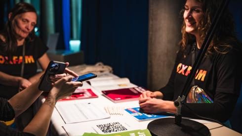 Zwei Oxfam-Mitarbeitende in schwarzen T-Shirts mit buntem Oxfam-Logo stehen um einen Info-Tisch und sprechen mit einer Person, die Infos zu unserer Petition erhalten will. Von dieser Person sind in der linken Ecke des Fotos nur die Arme und Hände sichtbar, sie macht mit ihrem Handy ein Foto von einem QR-Code.
