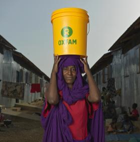 Eine Frau scheut in die KAmera. Auf dem Kopf trägt sie einen Wassereimer mit der Aufschrift Oxfam.